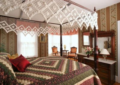 Prince of Wales guestroom with queen four poster bed topped with a handmade crocheted canopy. Two antique chairs in the bright five window bay. Antique dresser and original sink vanity.