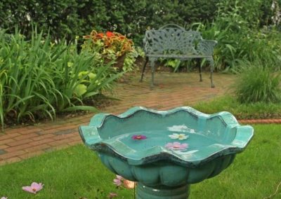 Closeup photo of a blue porcelain birdbath with purple and white daises floating in it. Lush gardens and iron settee in the background.