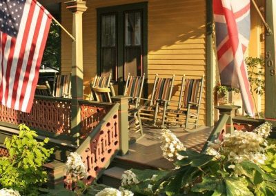 A close-up photo of the Prince Albert Hall porch on a very sunny day with rockers lining the porch and the American and British flags flying. White oakleaf hydrangea in the foreground.