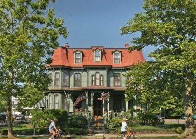 Photo taken of the front of The Queen Victoria in the summer. Two people are riding their bicycles by the house.